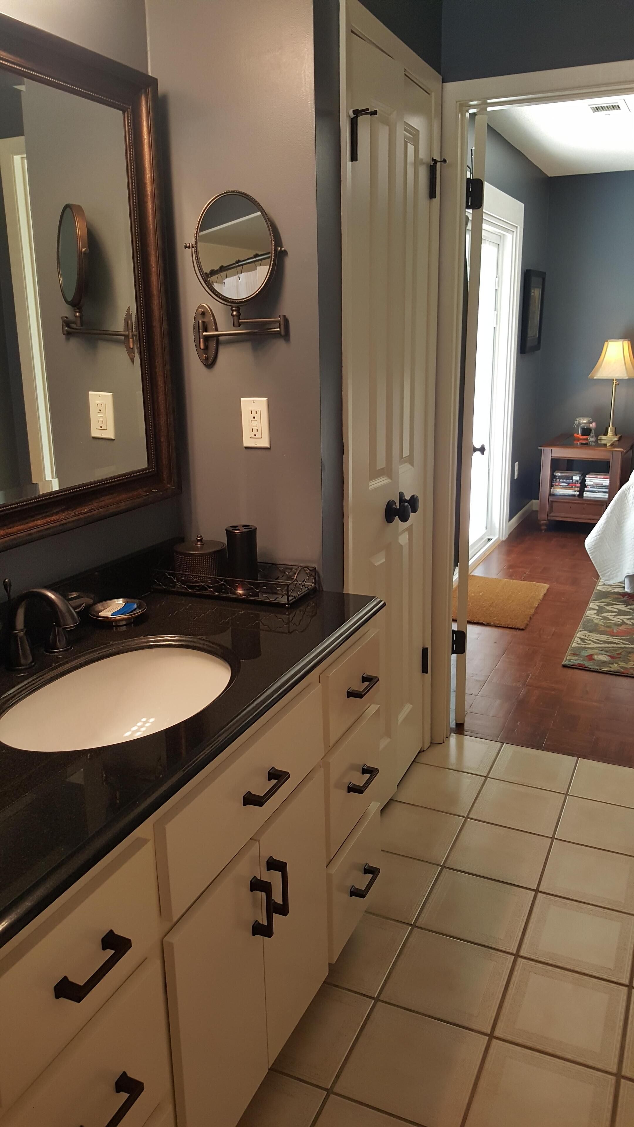 Une salle de bain avec un grand miroir, un lavabo et un meuble-lavabo avec des tiroirs. Une porte donne accès à une autre pièce.