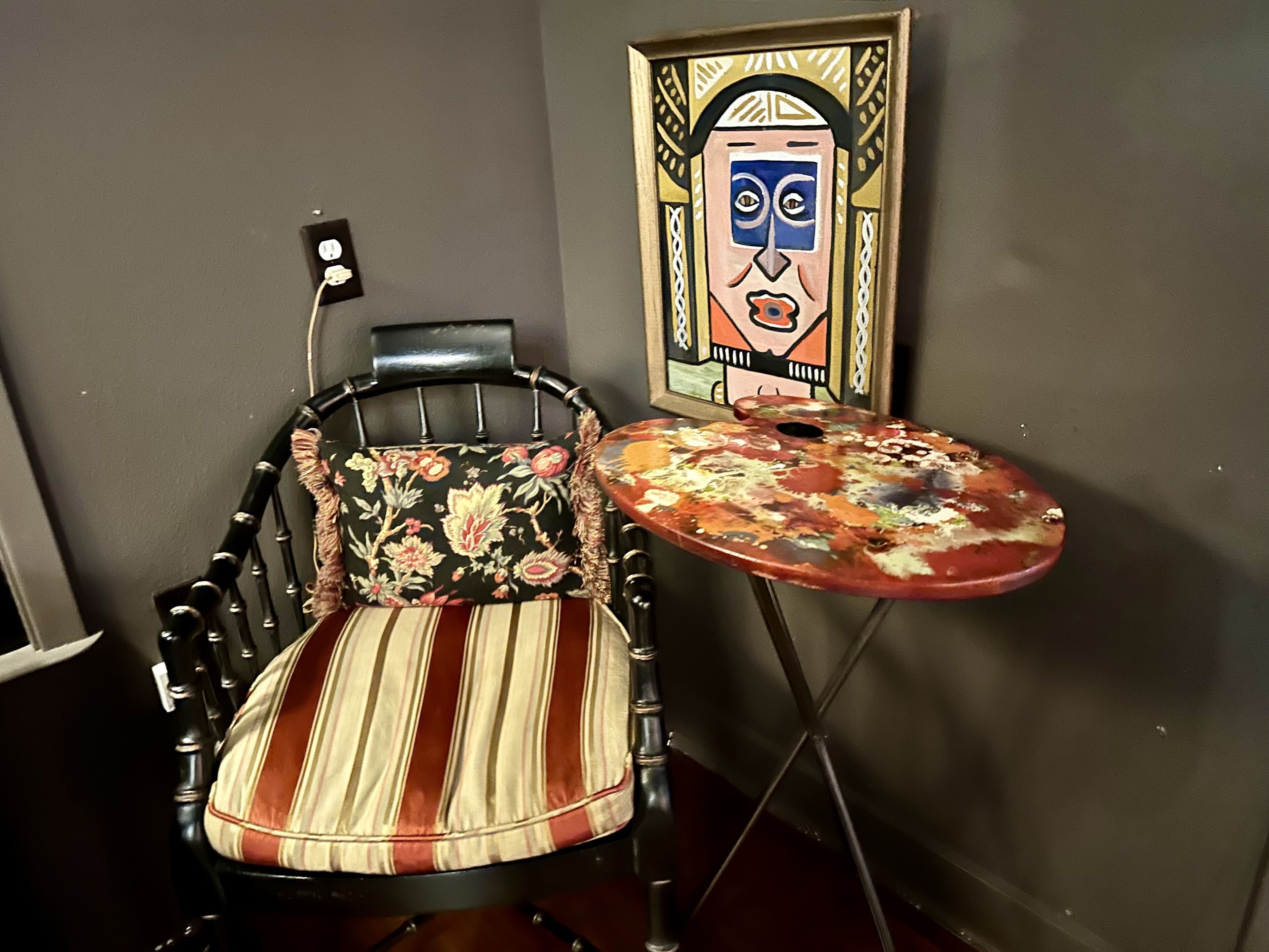 A chair with a floral cushion and a striped seat, a small table with a colorful, abstract design, and a framed artwork on the wall.