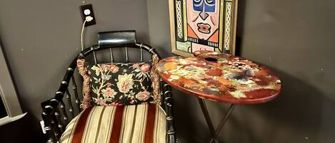 A chair with a floral cushion and a striped seat, a small table with a colorful, abstract design, and a framed artwork on the wall.