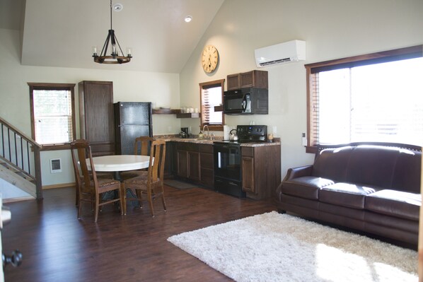 A spacious kitchen with wooden cabinets, a dining table, and chairs. There is a brown leather sofa and a white rug in the living area.