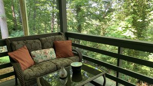 A wicker sofa with floral cushions on a glass-top table, set on a wooden deck with a view of a forest.