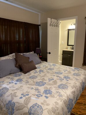 A bedroom with a floral bedspread, a wooden headboard, a nightstand with a lamp, and a door leading to a bathroom with a mirror and sink. - Feel Right At Home In This Spacious Retreat (Brevard)