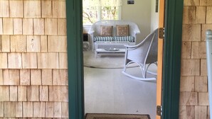Une façade en bois avec une porte verte donnant sur un intérieur où se trouvent un canapé blanc et un fauteuil à bascule.