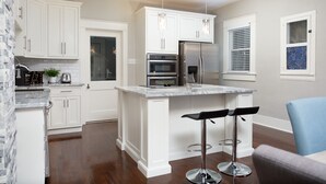 Eine moderne Küche mit weißen Schränken, einem Edelstahl-Kühlschrank, einem Backofen und einer Marmorarbeitsplatte. Zwei Barhocker und ein Fenster mit Jalousien sind ebenfalls vorhanden.