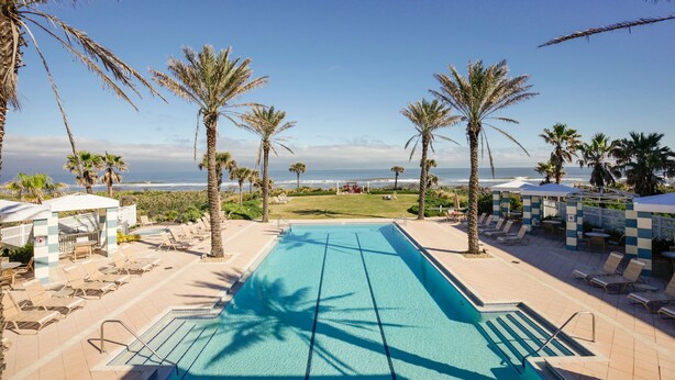 A pool area with lounge chairs and umbrellas, palm trees, and a view of the ocean. - Beachy Keen Platinum Property with Unbeatable View!   (Palm Coast)