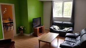 Un salon avec un mur vert, une table en bois, un canapé en cuir, un téléviseur posé sur un meuble en bois et une fenêtre avec des rideaux.