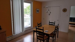 Une salle à manger avec une table et des chaises en bois, une fenêtre avec des rideaux blancs, une porte et une horloge murale.