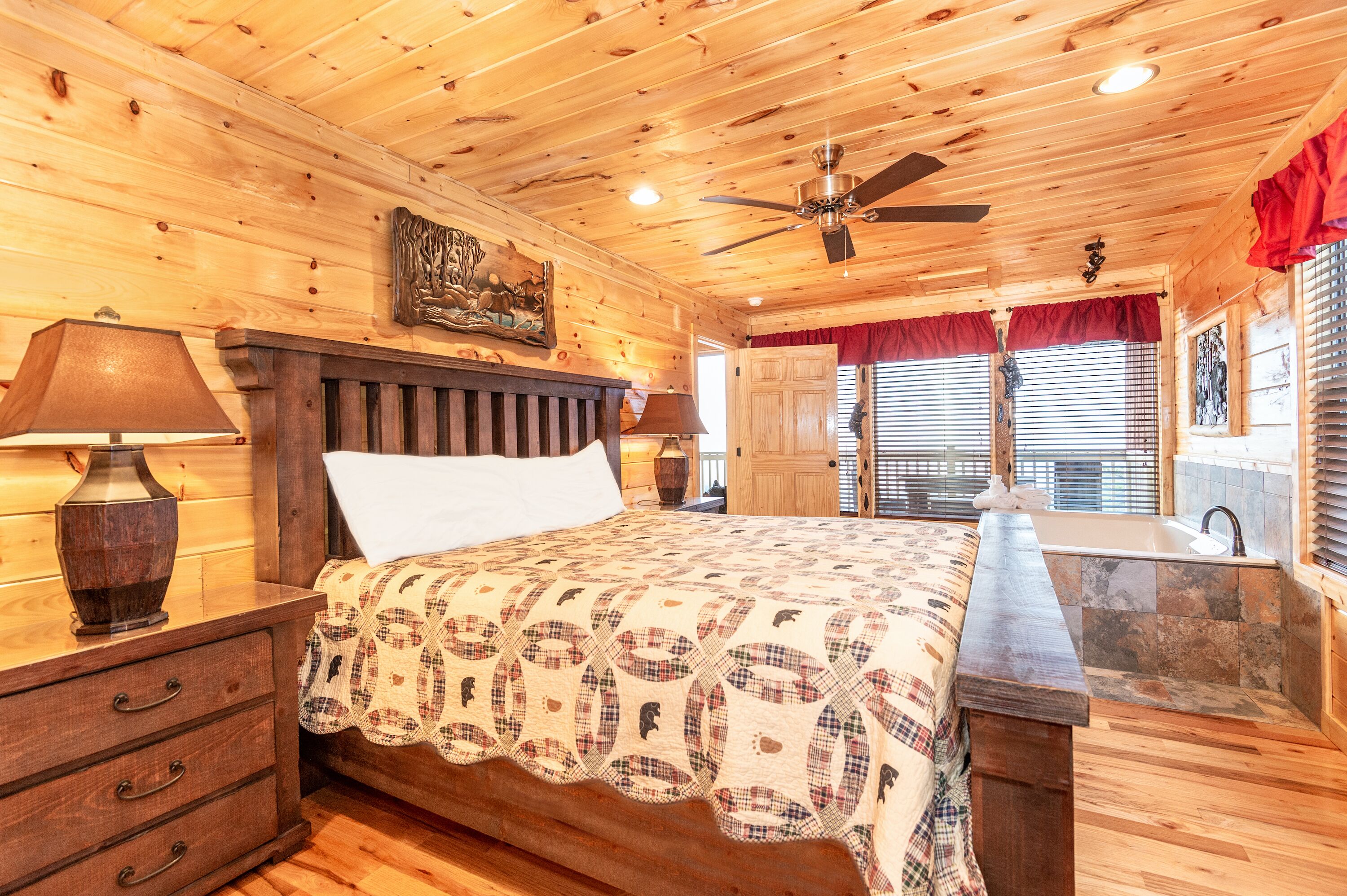 Une chambre dans une cabane en bois, comprenant un lit, deux lampes de chevet, un ventilateur de plafond, une fenêtre avec des stores et une salle de bain avec un lavabo et une douche.