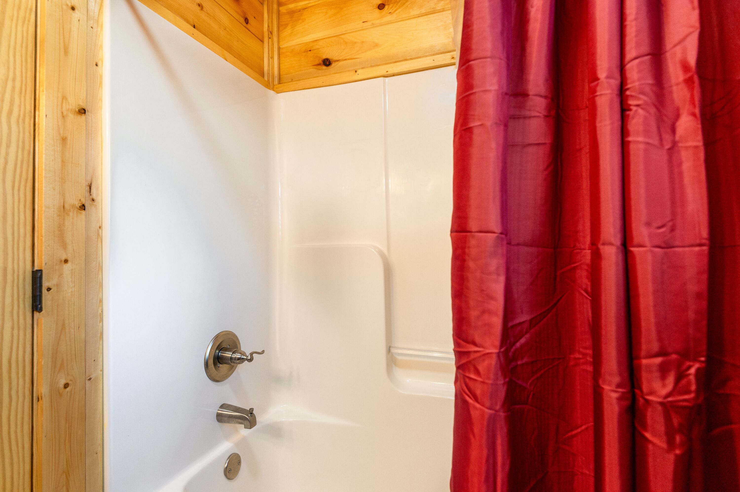 A corner of a room with a wooden ceiling, a white bathtub, and a red shower curtain.
