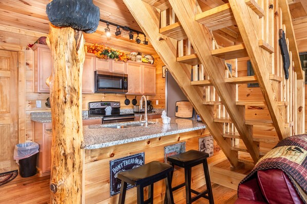 A wooden cabin interior with a kitchen area, a staircase, and a bar stool.