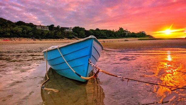 Ein blaues Boot liegt an einem Steg am Strand, im Hintergrund ein Sonnenuntergang.