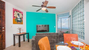 Ein modernes Wohnzimmer mit einer türkisfarbenen Akzentwand, einem grauen Sofa, orangefarbenen Sesseln, einem Esstisch für zwei Personen und einem Deckenventilator.