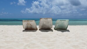 Drei verwitterte Betonstühle an einem sandigen Strand, im Hintergrund ein klarer, blauer Himmel und ein türkisblaues Meer.