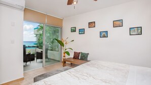 Ein Schlafzimmer mit einem Bett, einem Deckenventilator, einer Bank, einer Topfpflanze und Blick auf das Meer durch eine Schiebetür.