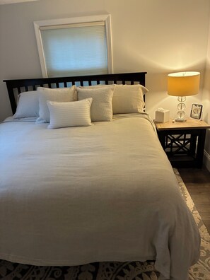 A neatly made bed with a white comforter and pillows, a bedside table with a lamp and a tissue box, and a window with blinds.