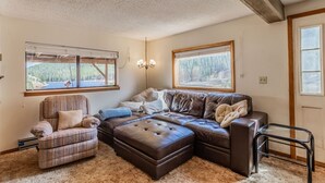 A cozy living room with a sectional sofa, a recliner, and a large window offering a view of a forested area. - Fawn Haven on the Lake (Bailey)