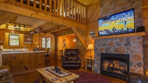 A cozy cabin interior with a stone fireplace, wooden furniture, and a television displaying a sports event. - Gorgeous, Secluded, SuperFast WiFi, NFL Sunday, Arcade, Gated, Fishing & 2 Pools (Sevierville)