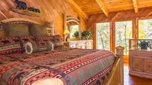 A log cabin bedroom with a large bed, wooden walls, and a view of trees through the windows. - Gorgeous, Secluded, SuperFast WiFi, NFL Sunday, Arcade, Gated, Fishing & 2 Pools (Sevierville)