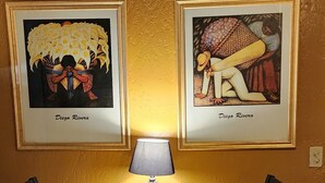 A dining area with a round table, two chairs, a lamp, and two framed artworks on the wall.