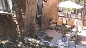 A small brick cabin with a chimney, a door, and a window. There is a patio area with a table, chairs, and potted plants.