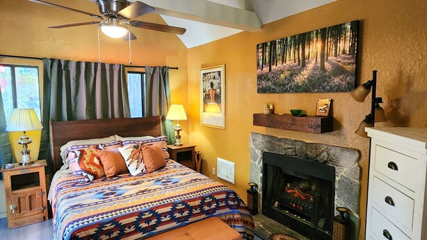 A bedroom with a bed, a fireplace, a ceiling fan, and a painting on the wall.