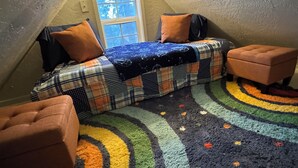 A cozy attic bedroom with a sloped ceiling, a window, a bed with plaid bedding, two ottomans, and a multicolored rug.