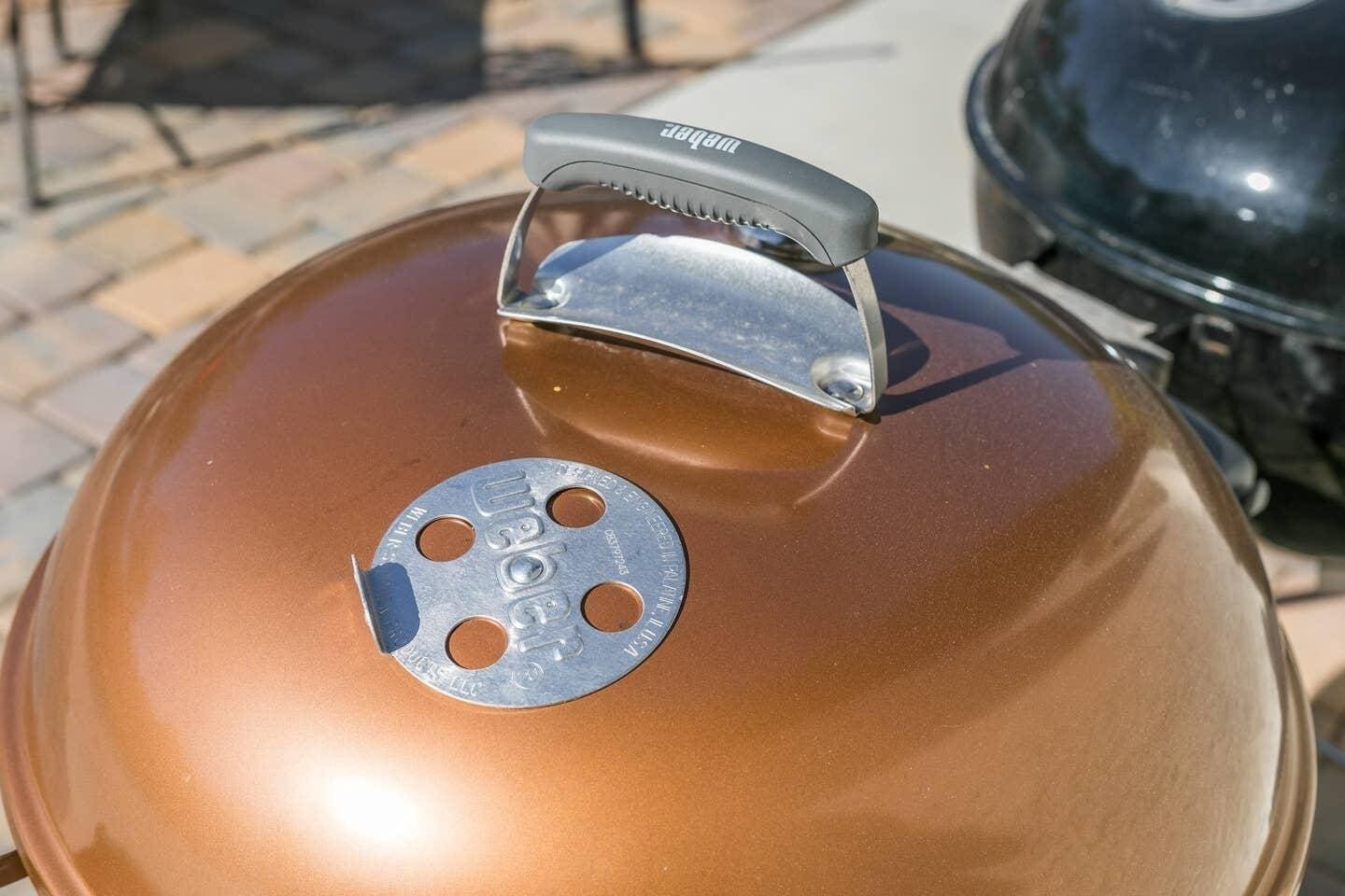 A round, metallic lid with a handle, a vent, and a label on a barbecue grill.