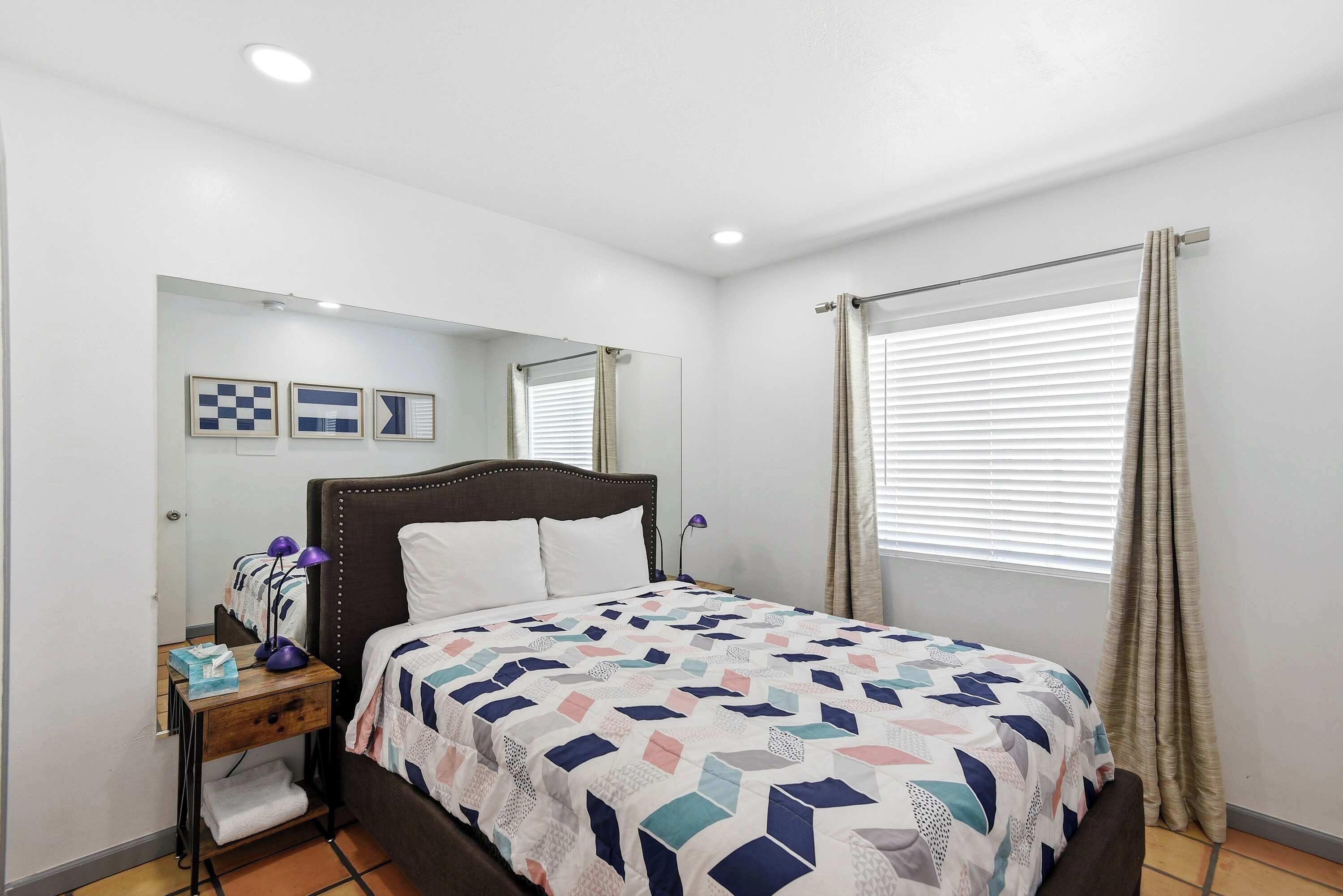 Un dormitorio con cama, mesita de noche, lámpara, espejo y ventana con cortinas.