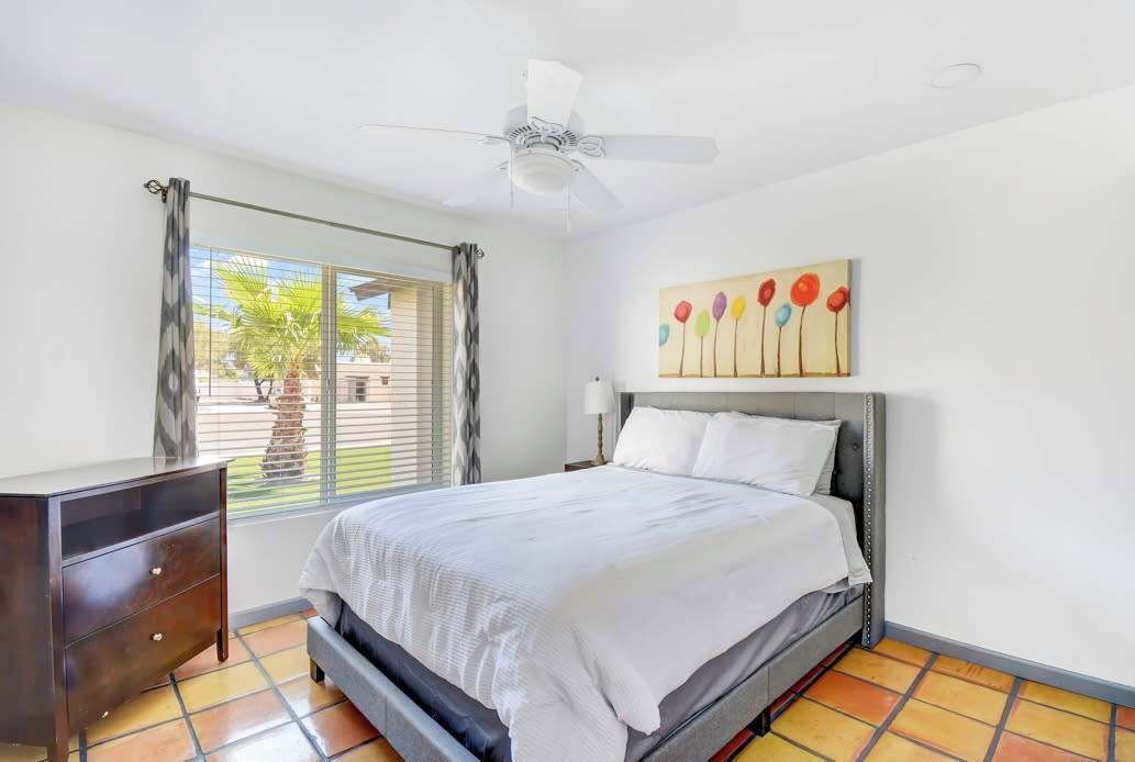 A bedroom with a bed, a ceiling fan, a window with curtains, a bedside table, and a painting on the wall.