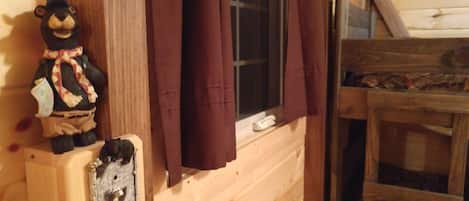 A rustic cabin interior with a wooden wall, a window with brown curtains, and a small bear figurine on a shelf.