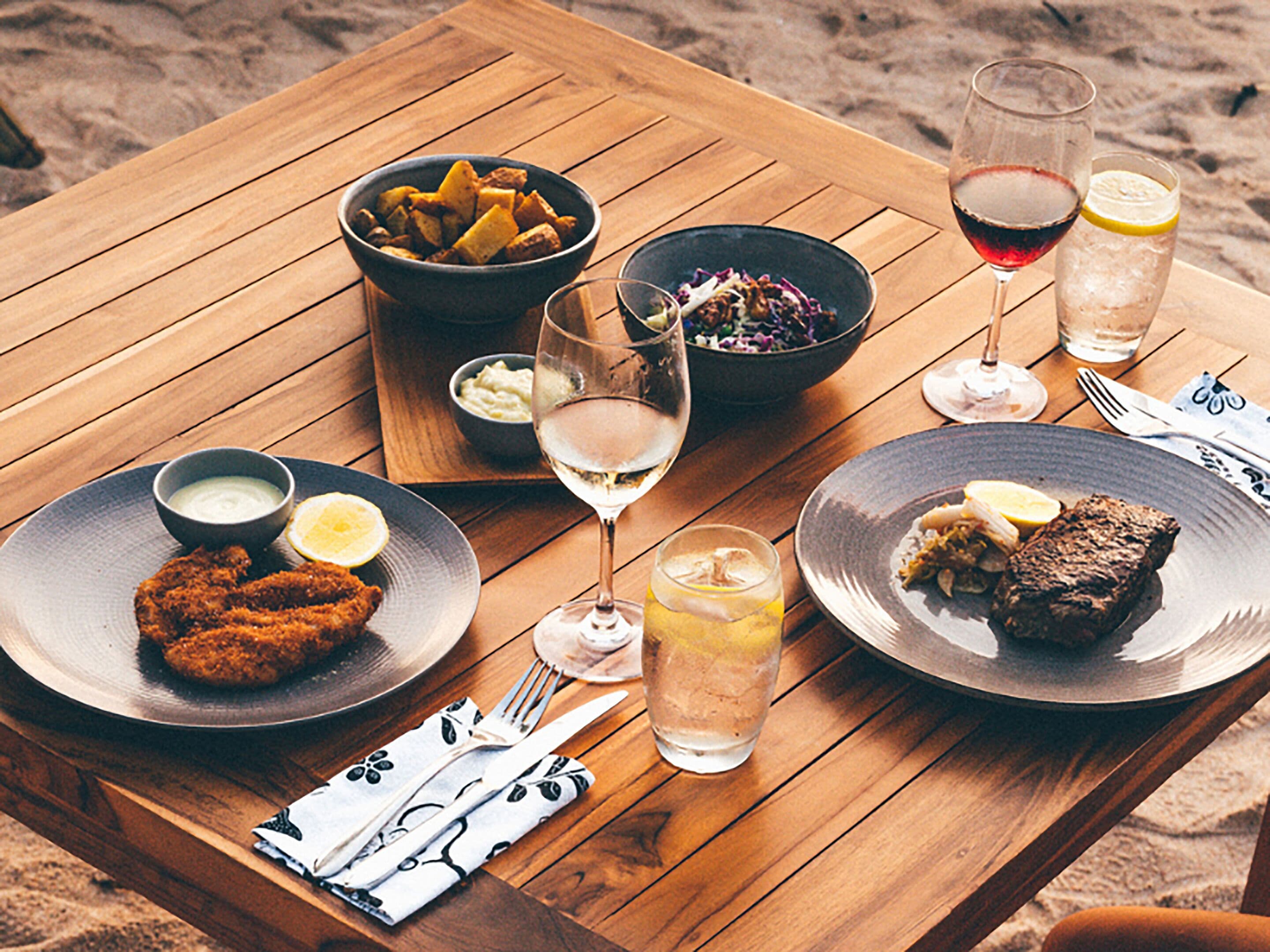 A wooden table with various dishes, including fried items, salad, and a steak, along with glasses of wine and water.