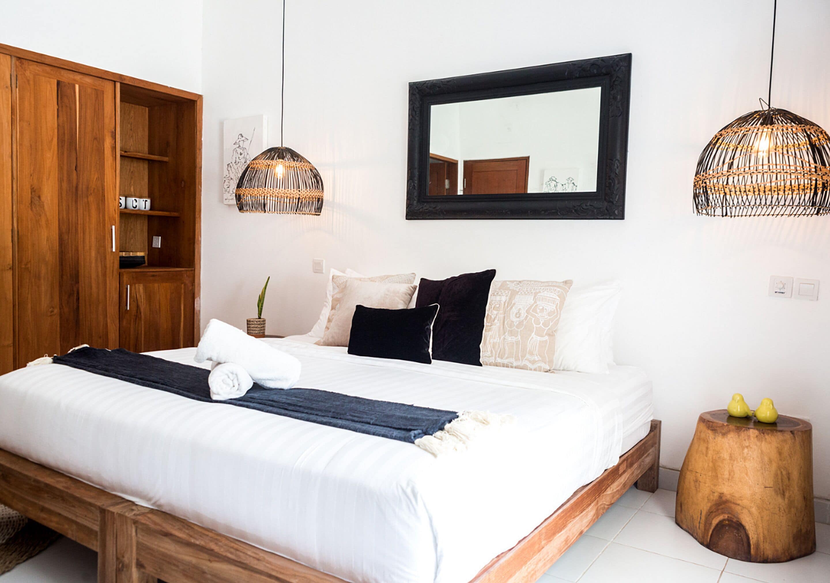Un lit bien fait, avec une tête de lit en bois, un grand miroir au-dessus et deux suspensions lumineuses en osier.
