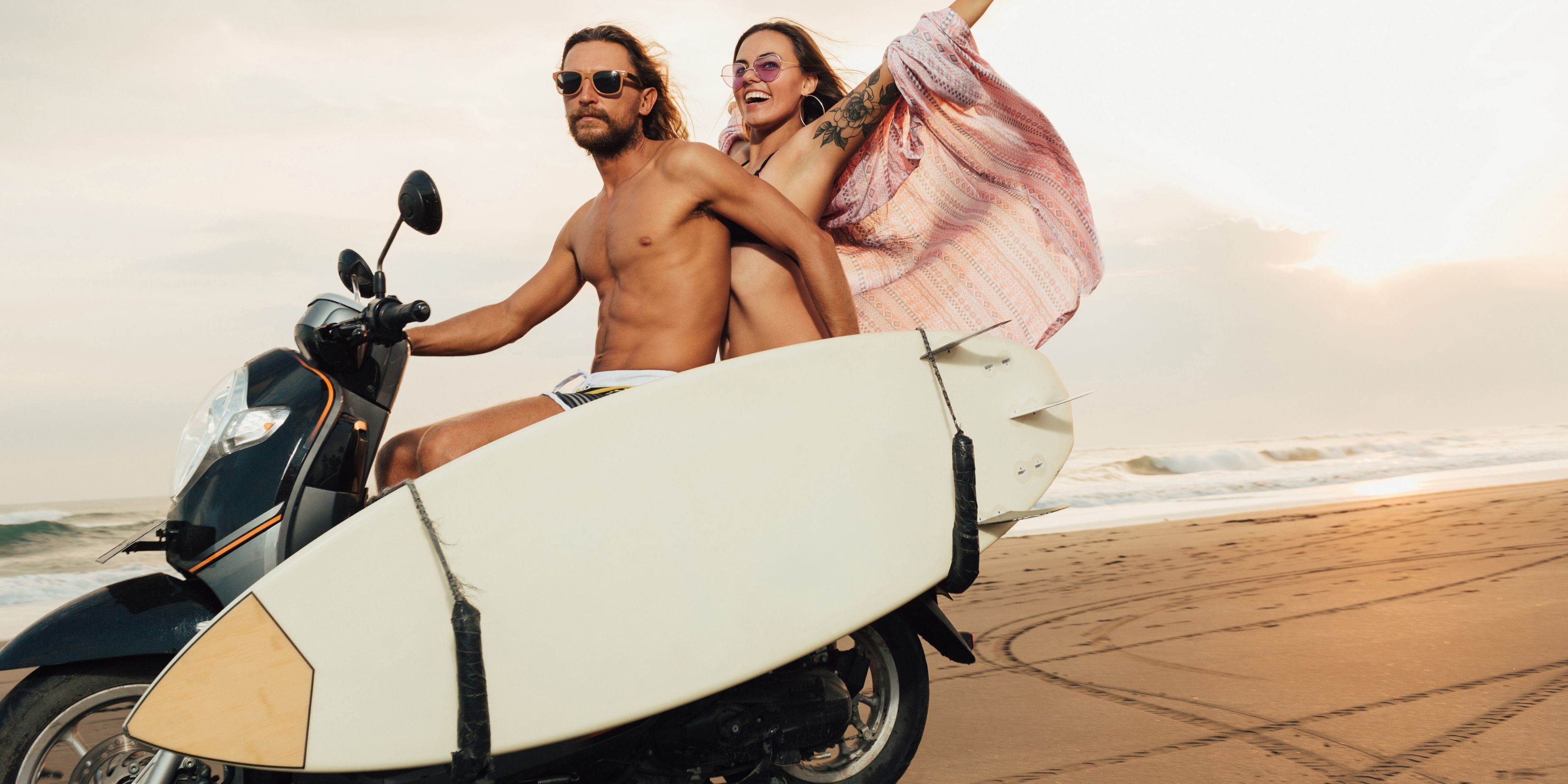 A person riding a scooter with a surfboard attached, on a beach.
