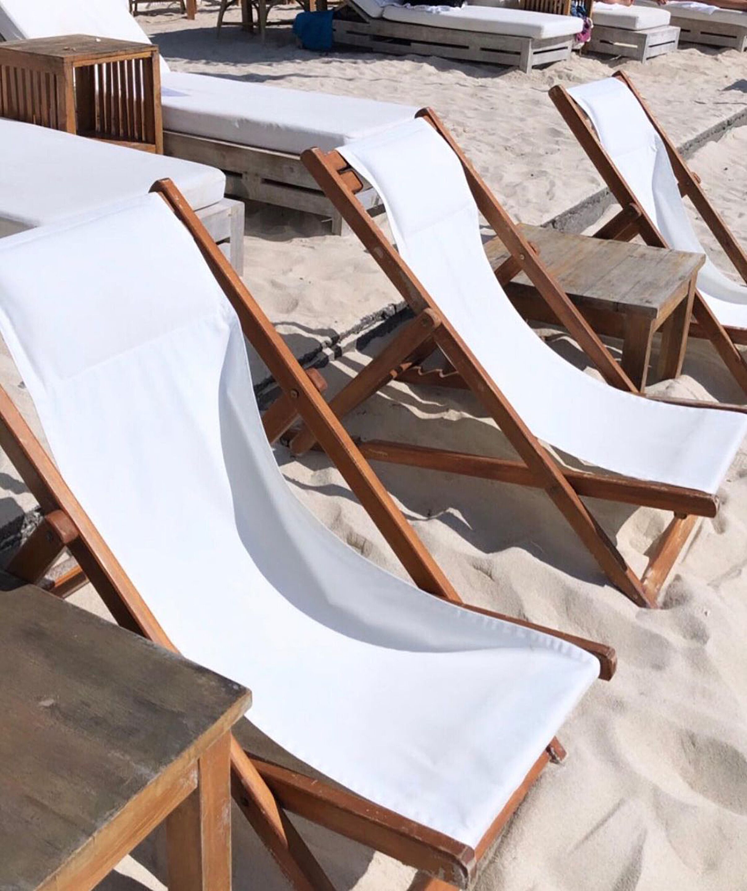 A row of wooden lounge chairs with white cushions on a sandy beach.