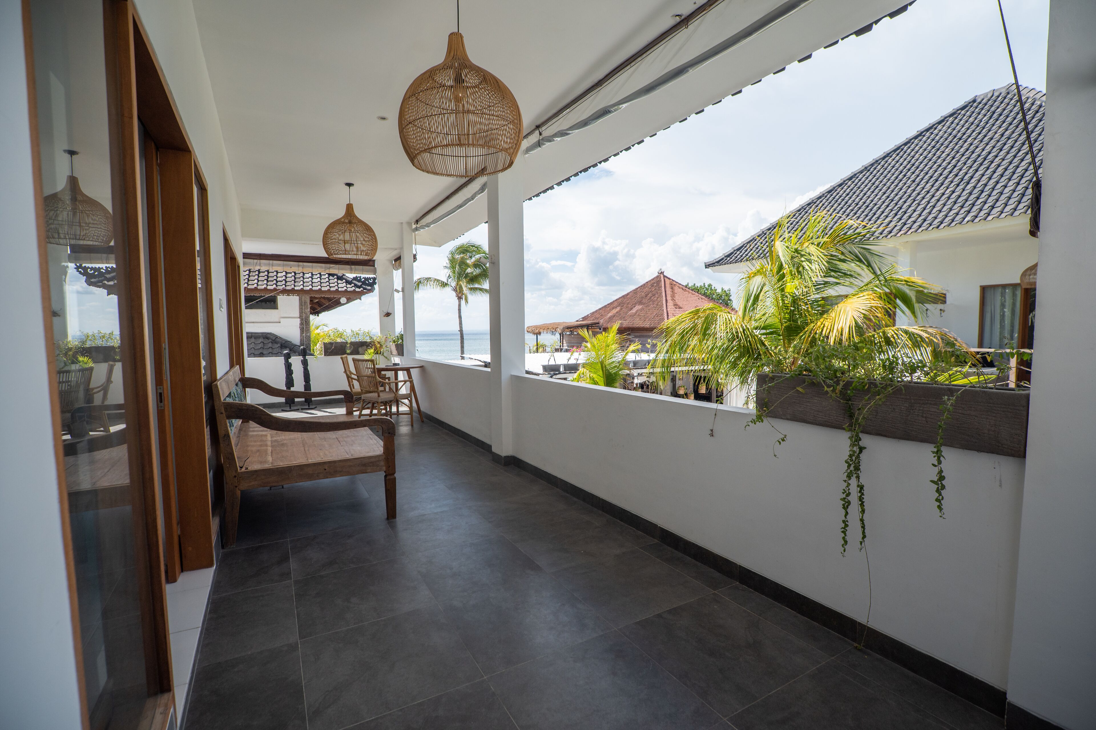 A balcony with a wooden bench and hanging wicker lamps, overlooking a coastal area with palm trees and a traditional house.