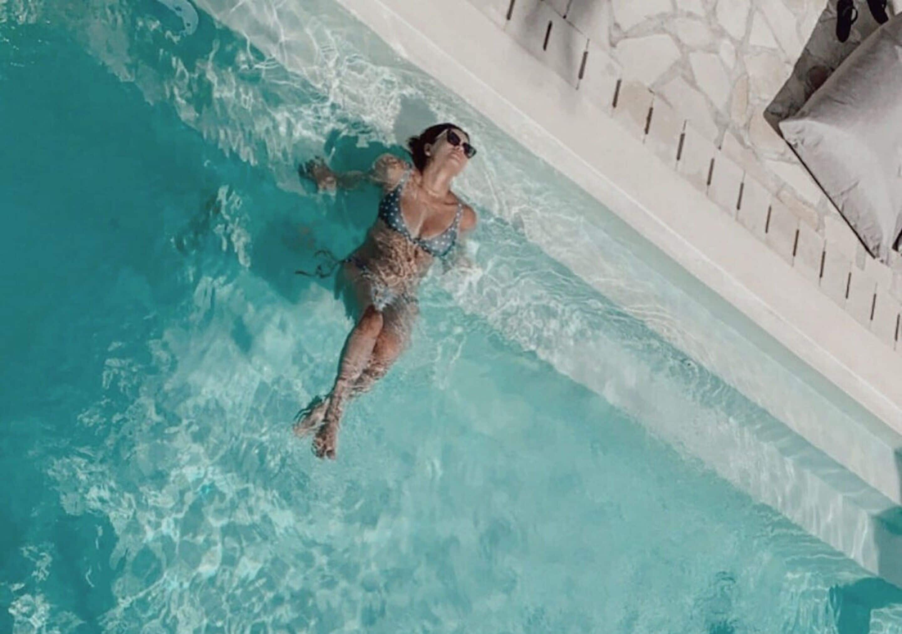 Une personne flotte dans une piscine à l’eau claire et bleue, bordée d’un mur de pierre et ornée de piliers blancs.