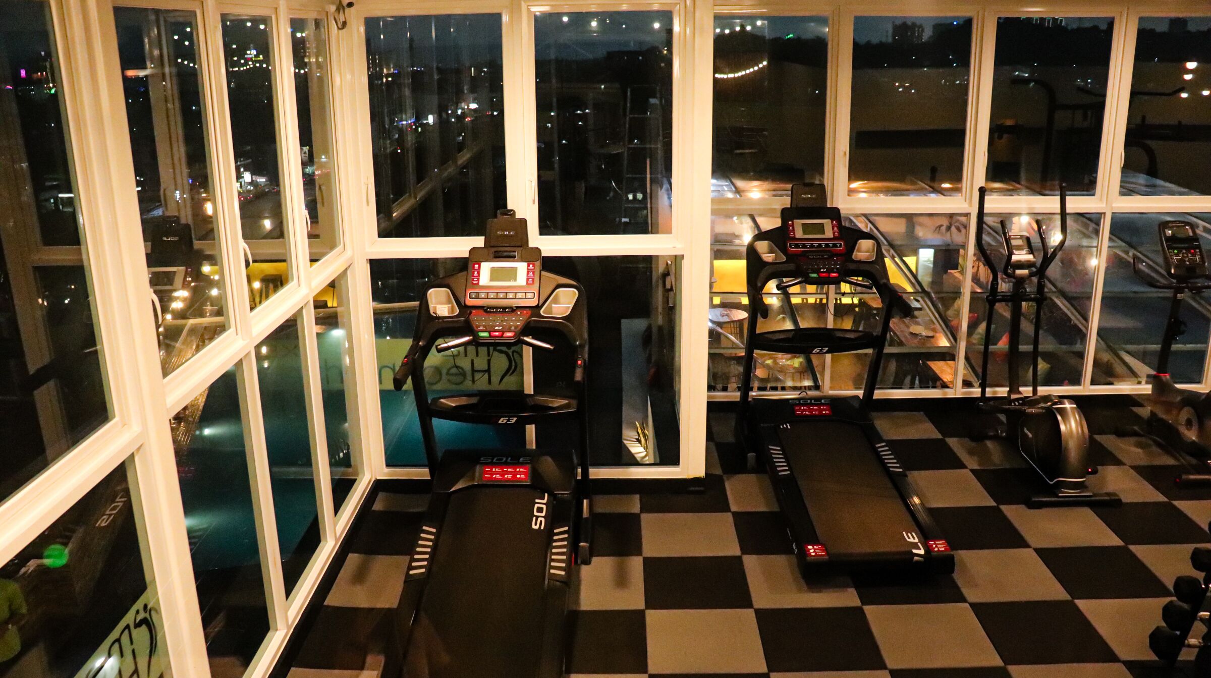Un gimnasio con cintas de correr y equipamiento de ejercicio, amplias ventanas y suelo a cuadros.