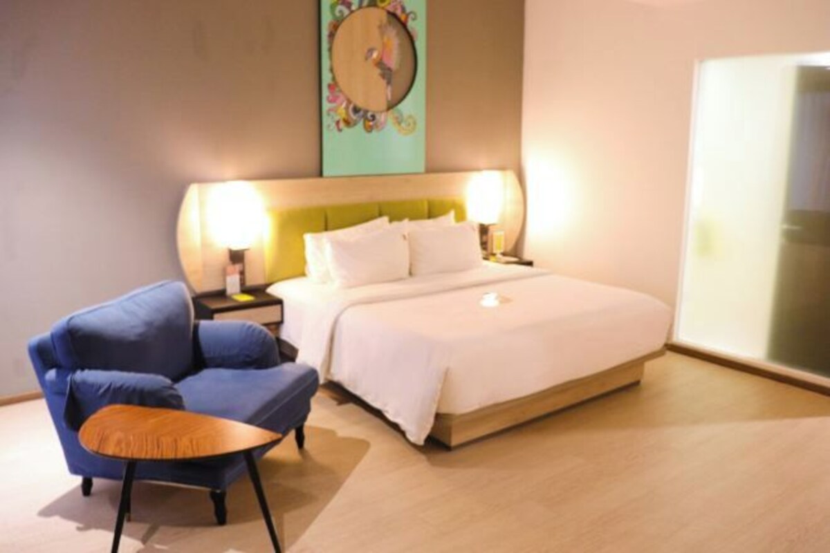 A hotel room with a bed, bedside lamps, a blue armchair, a small wooden table, and a wall art piece.