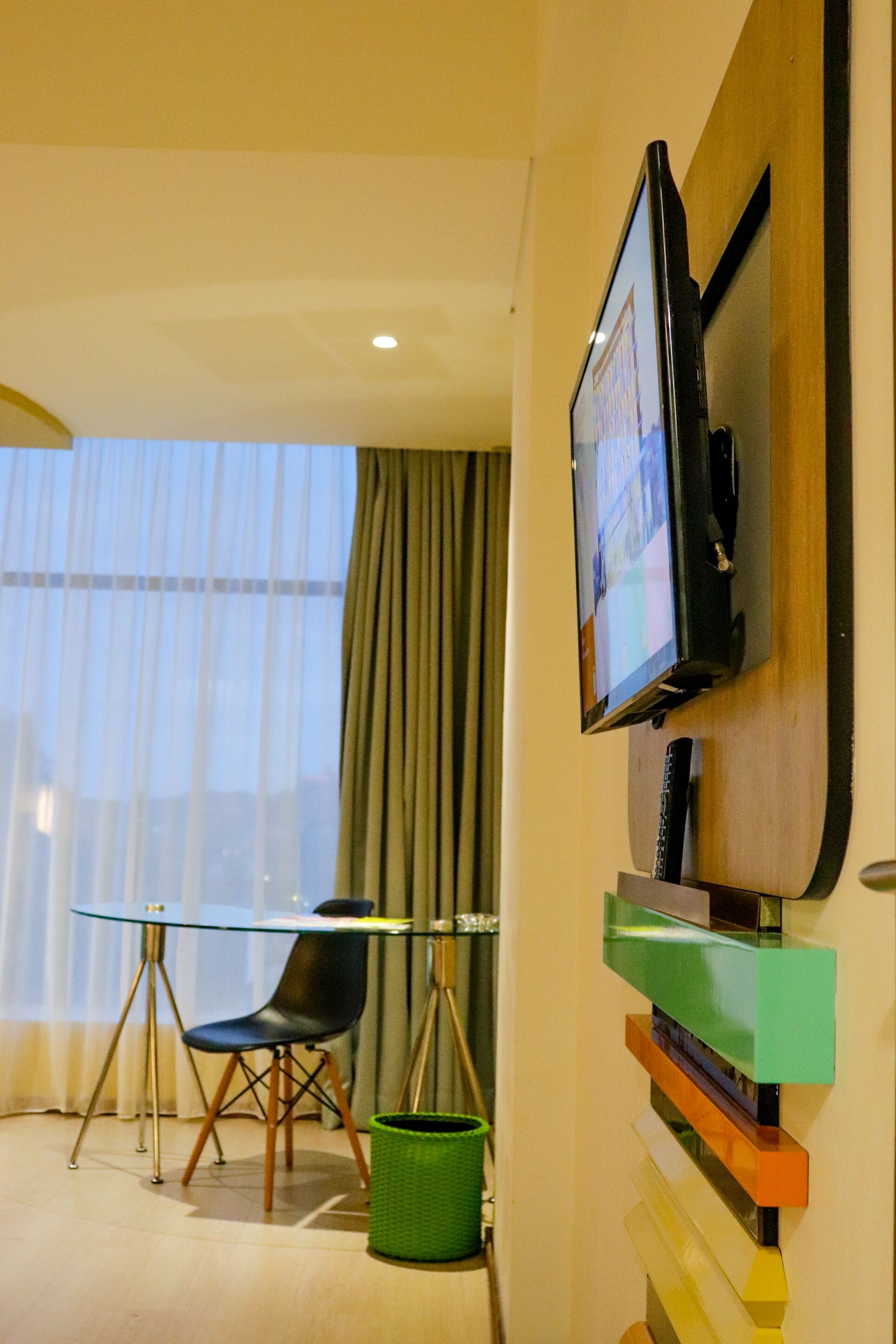 A hotel room with a glass table, a black chair, a green trash bin, and a television mounted on the wall.