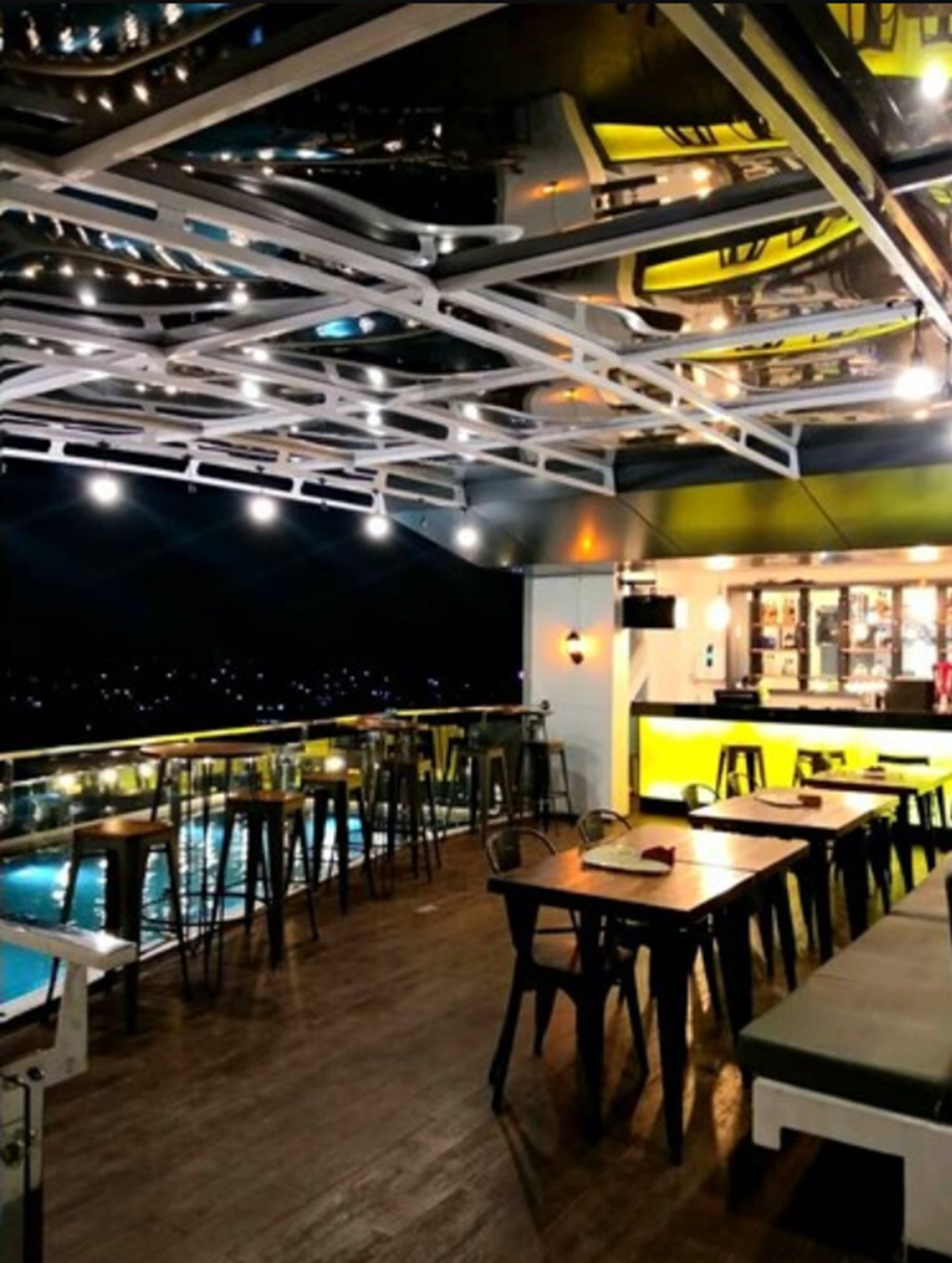A rooftop bar with a pool, wooden tables, and chairs, featuring a modern ceiling design and a bar area with a counter and stools.