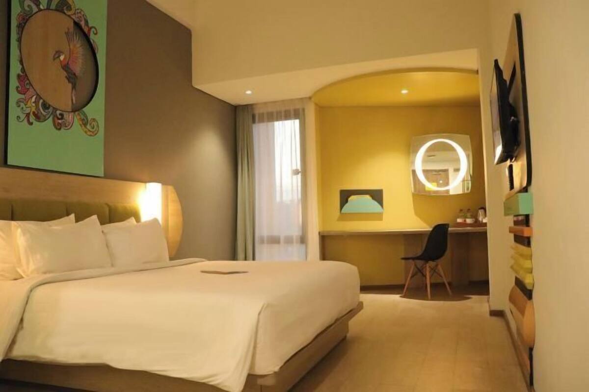 A hotel room with a bed, a desk, a chair, a round mirror, and a wall clock.