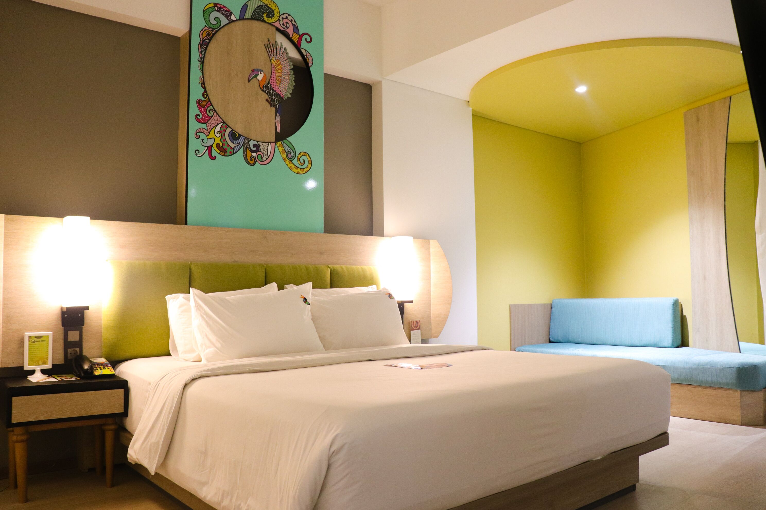 A hotel room with a large bed, a bedside table with a lamp, a small desk with a telephone, and a colorful wall decoration.