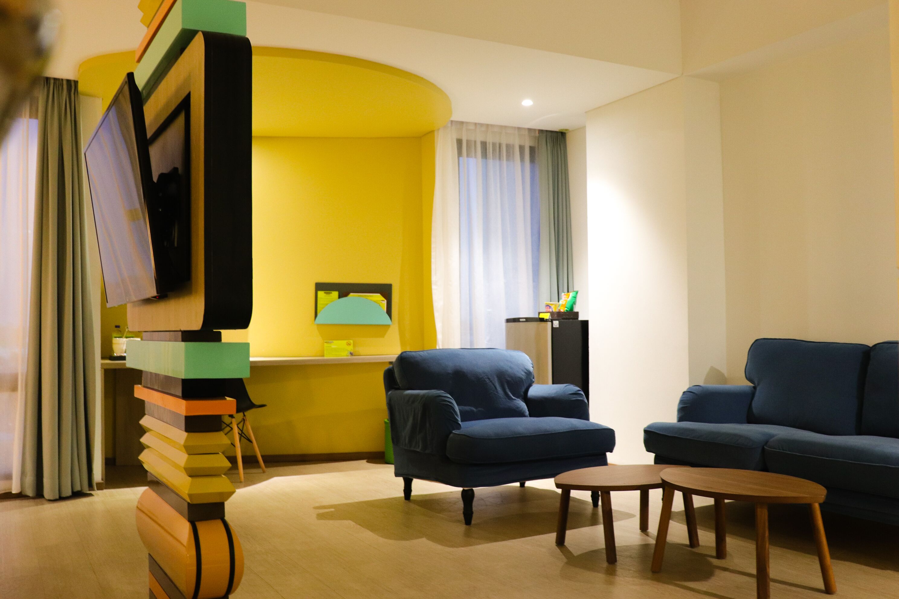 Un salon avec un canapé bleu, un fauteuil bleu et une petite table basse en bois. Un téléviseur est fixé au mur et une lampe se trouve sur une table d’appoint.