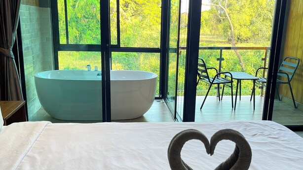 Ein Hotelzimmer mit einem großen Bett, einer herzförmig arrangierten Handtuchgarnitur, einer Badewanne und einem Balkon mit Blick auf Grünflächen.
