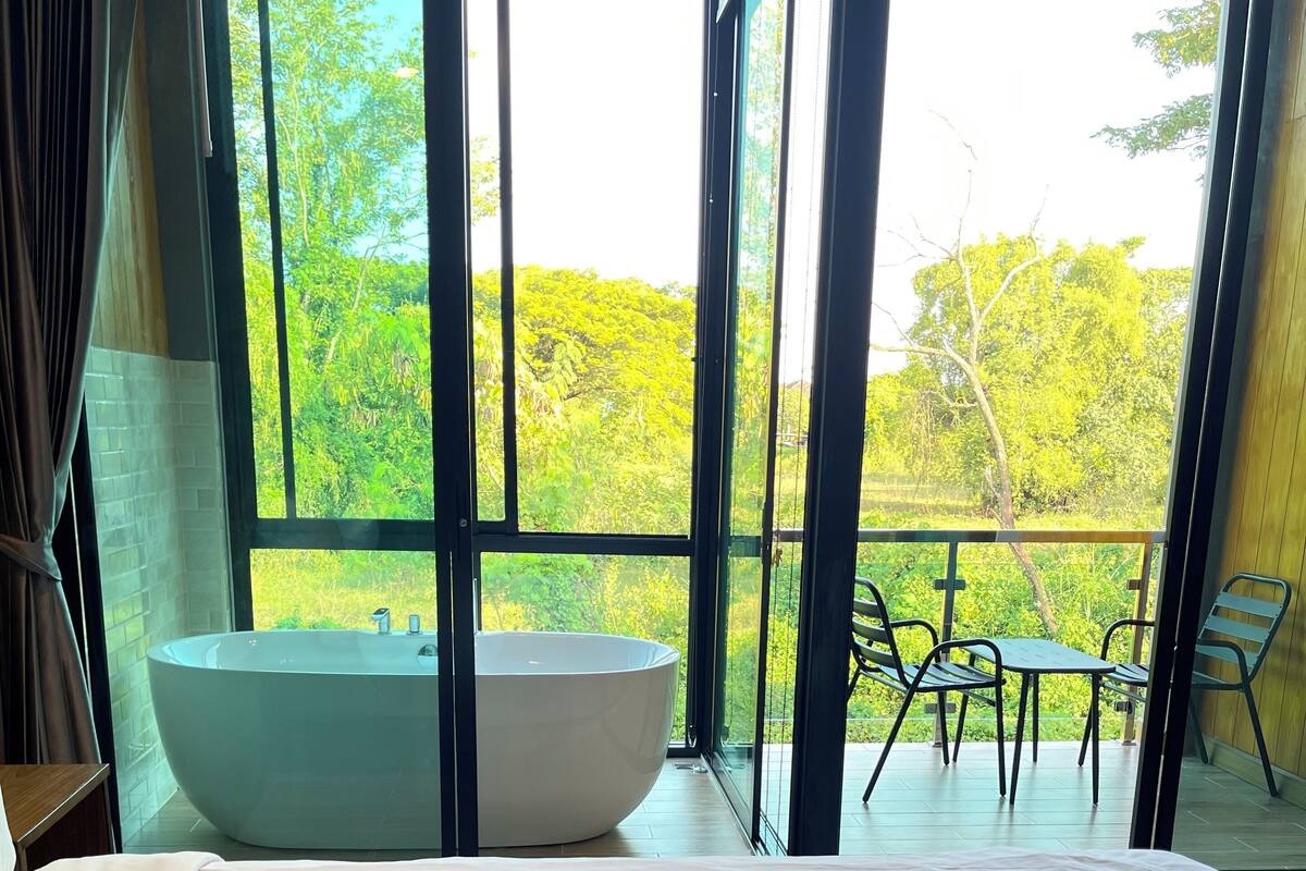 A hotel room with a large bed, a heart-shaped towel arrangement, a bathtub, and a balcony with a view of greenery.