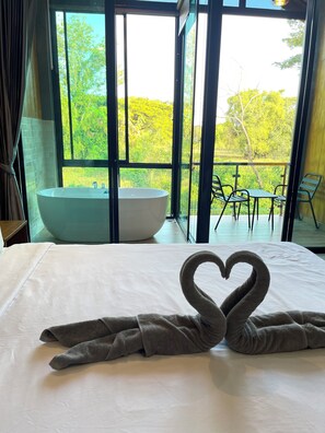 Habitación de hotel con una cama grande, una toalla dispuesta en forma de corazón, una bañera y un balcón con vistas a la vegetación.
