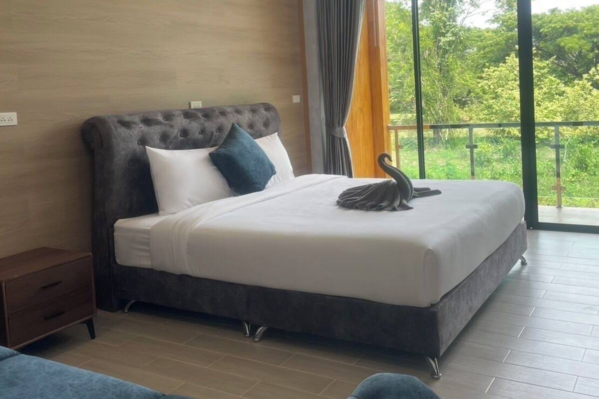 A modern hotel room with a large bed, a nightstand, a blue sofa, and a balcony with a view of greenery.