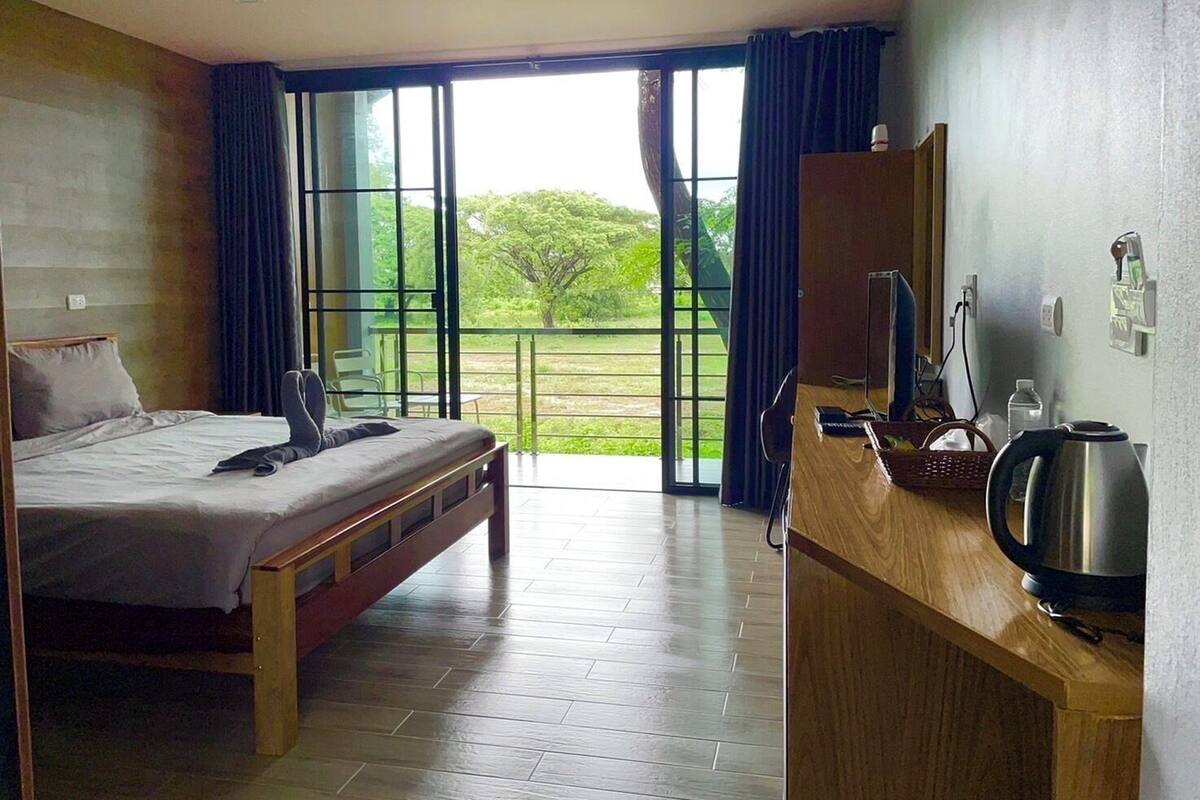 A hotel room with a bed, a desk with a kettle, and a view of greenery through large windows.