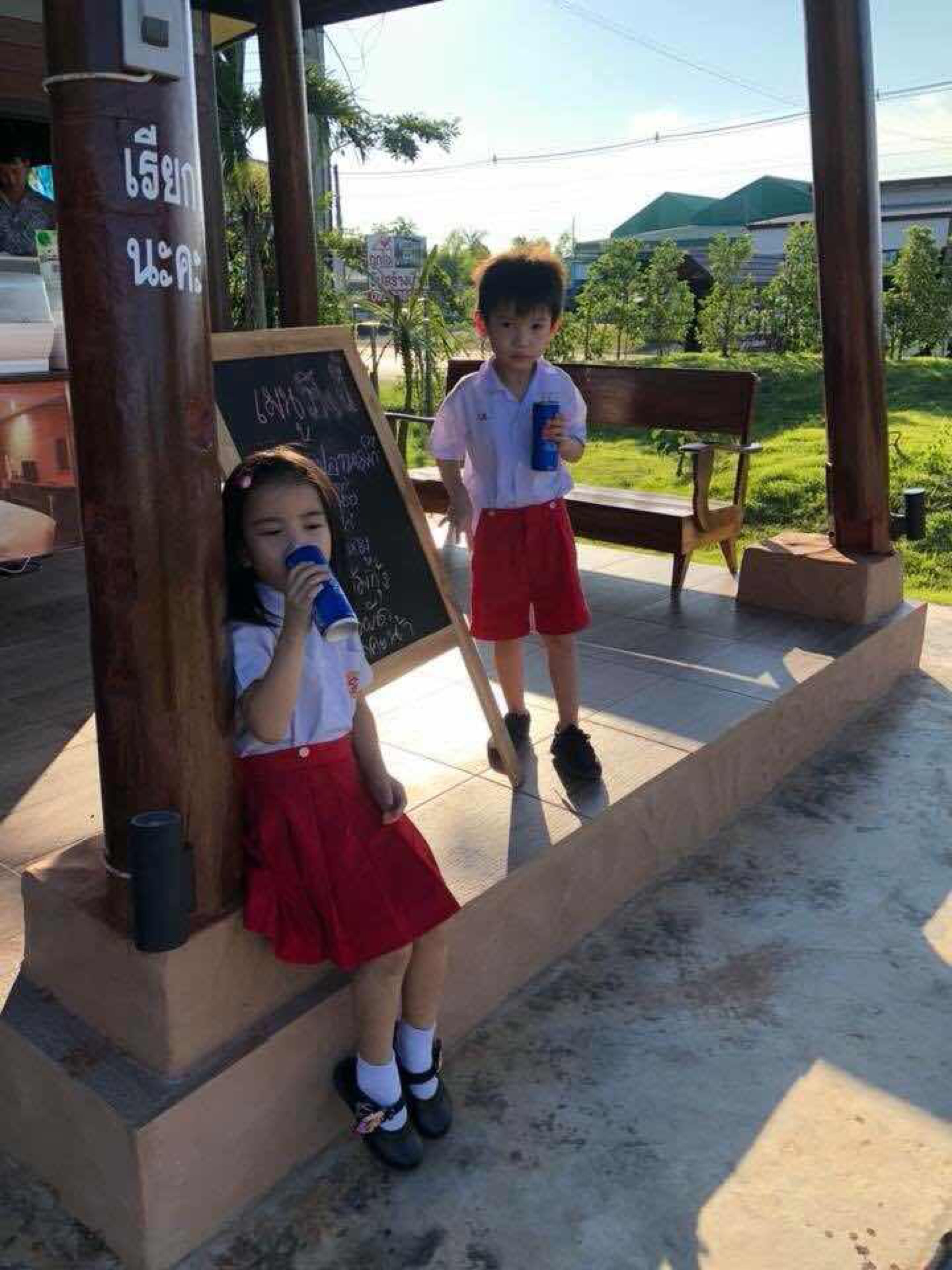Two children standing near a chalkboard in an outdoor setting with a bench and greenery in the background.