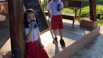 Two children standing near a chalkboard in an outdoor setting with a bench and greenery in the background.